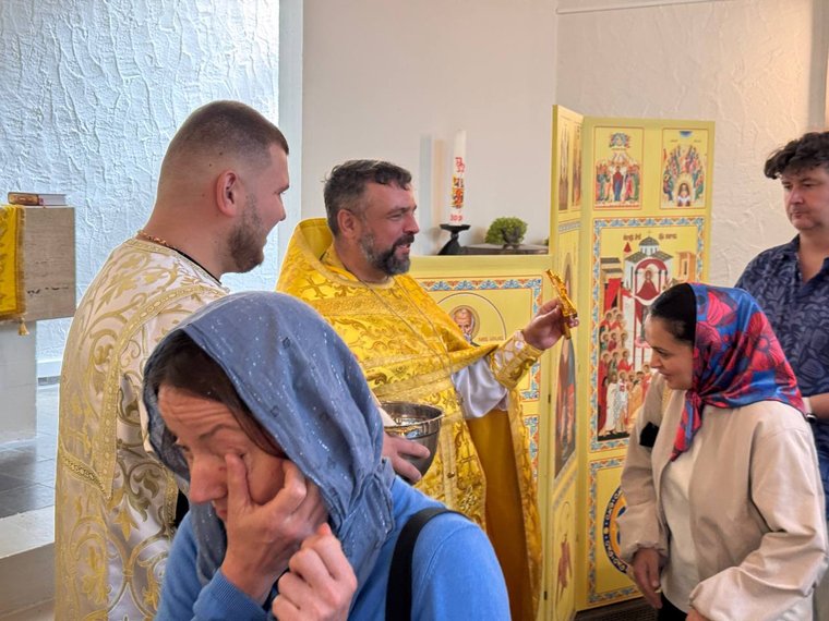 Парафіянка в блакитній хустці витирає краплі свяченої води, позаду священник у золотих ризах